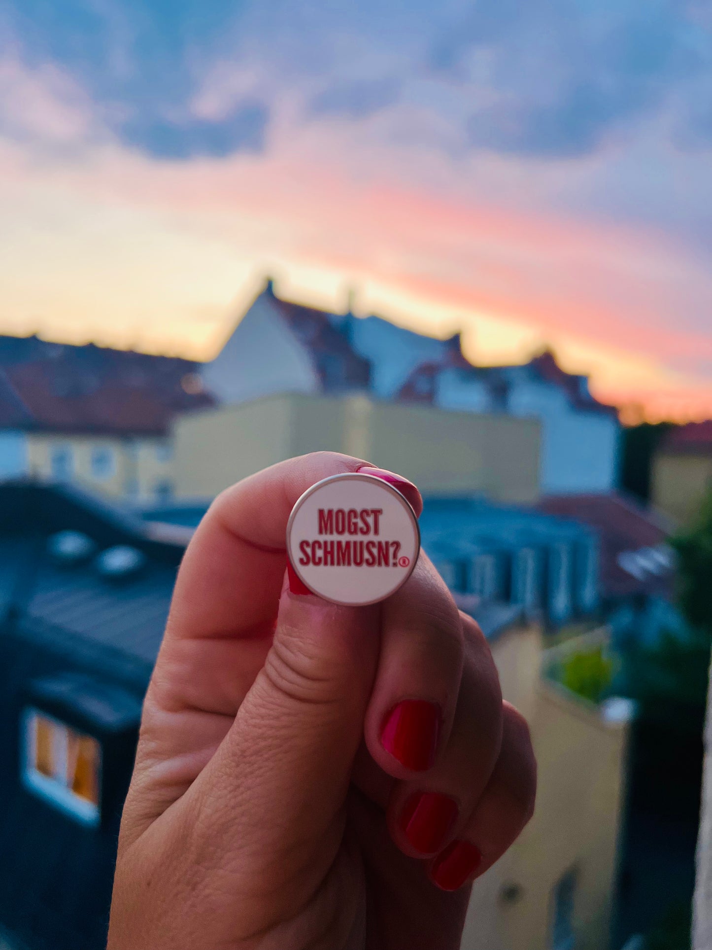 MOGST SCHMUSN?® PIN in Markenrot mit Premium Butterfly-Verschluss – wir in der Hand gegen den Abendhimmel gehalten