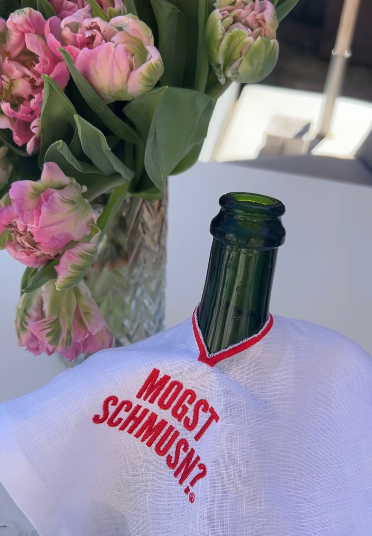 Weinflasche mit weißem MOGST SCHMUSN?® Weintuch aus Leinen und roter Stickerei, dekoriert mit Blumen im Hintergrund