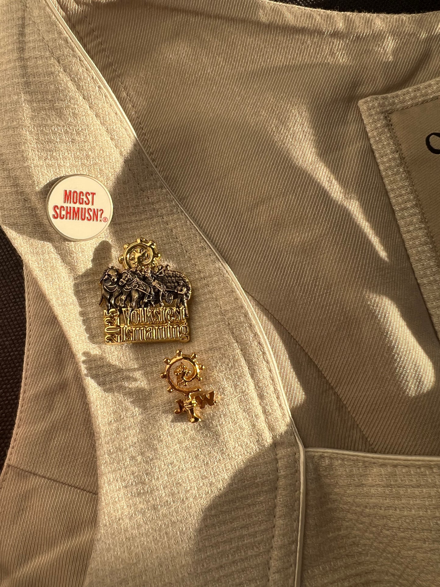 MOGST SCHMUSN?® PIN in Markenrot mit Premium Butterfly-Verschluss – angesteckt auf einem cremefarbenen Dirndl - auf dem Bild sind ebenfalls PINs vom Ismaninger Volksfest und die Augustiner Stammgastnadel