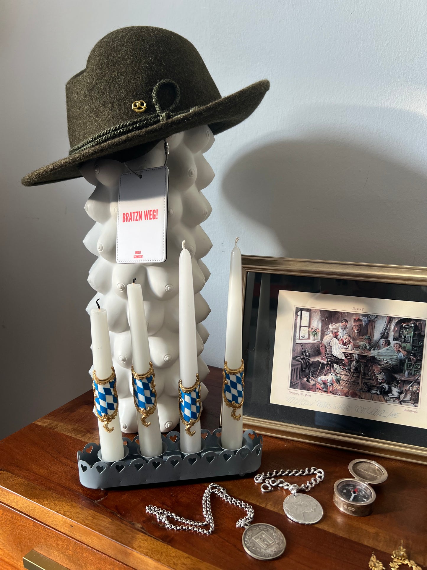 MOGST SCHMUSN?® Koffer- und Taschenanhänger mit rotem Schriftzug „BRATZN WEG!“ an einer weißen Designfigur mit braunem Trachtenhut, inszeniert auf einem Holztisch mit bayerischem Kerzenhalter, weiß-blauen Kerzen, Schmuck und gerahmter Illustration im Hintergrund.