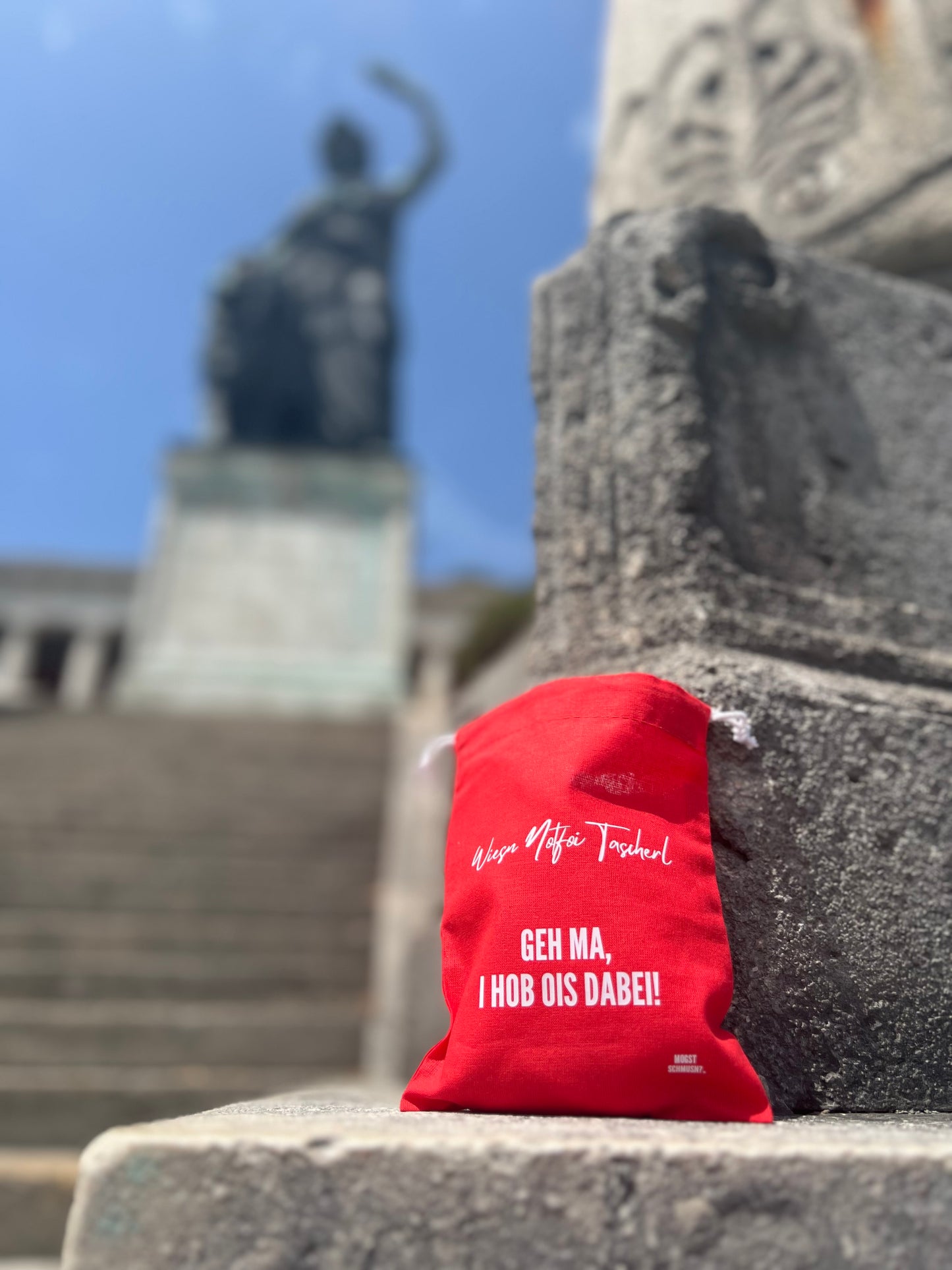 Rotes MOGST SCHMUSN?® Wiesn Notfoi Tascherl mit weißer Aufschrift vor der Bavaria-Statue in München