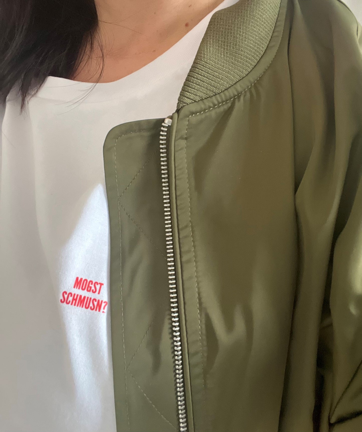 Ein Model trägt unter ihrer grünen Bombajacke, das weißes Unisex-T-Shirt mit kleinem roten MOGST SCHMUSN?® Aufdruck auf der Brust (links). Normal geschnitten, nachhaltig produziert aus Bio-Baumwolle.