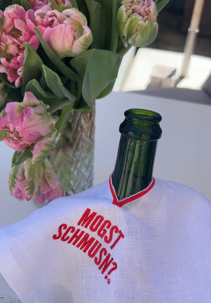 Weinflasche mit weißem MOGST SCHMUSN?® Weintuch aus Leinen und roter Stickerei, dekoriert mit Blumen im Hintergrund