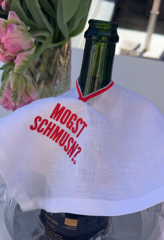 Weinflasche mit weißem MOGST SCHMUSN?® Weintuch aus Leinen und roter Stickerei, dekoriert mit Blumen im Hintergrund