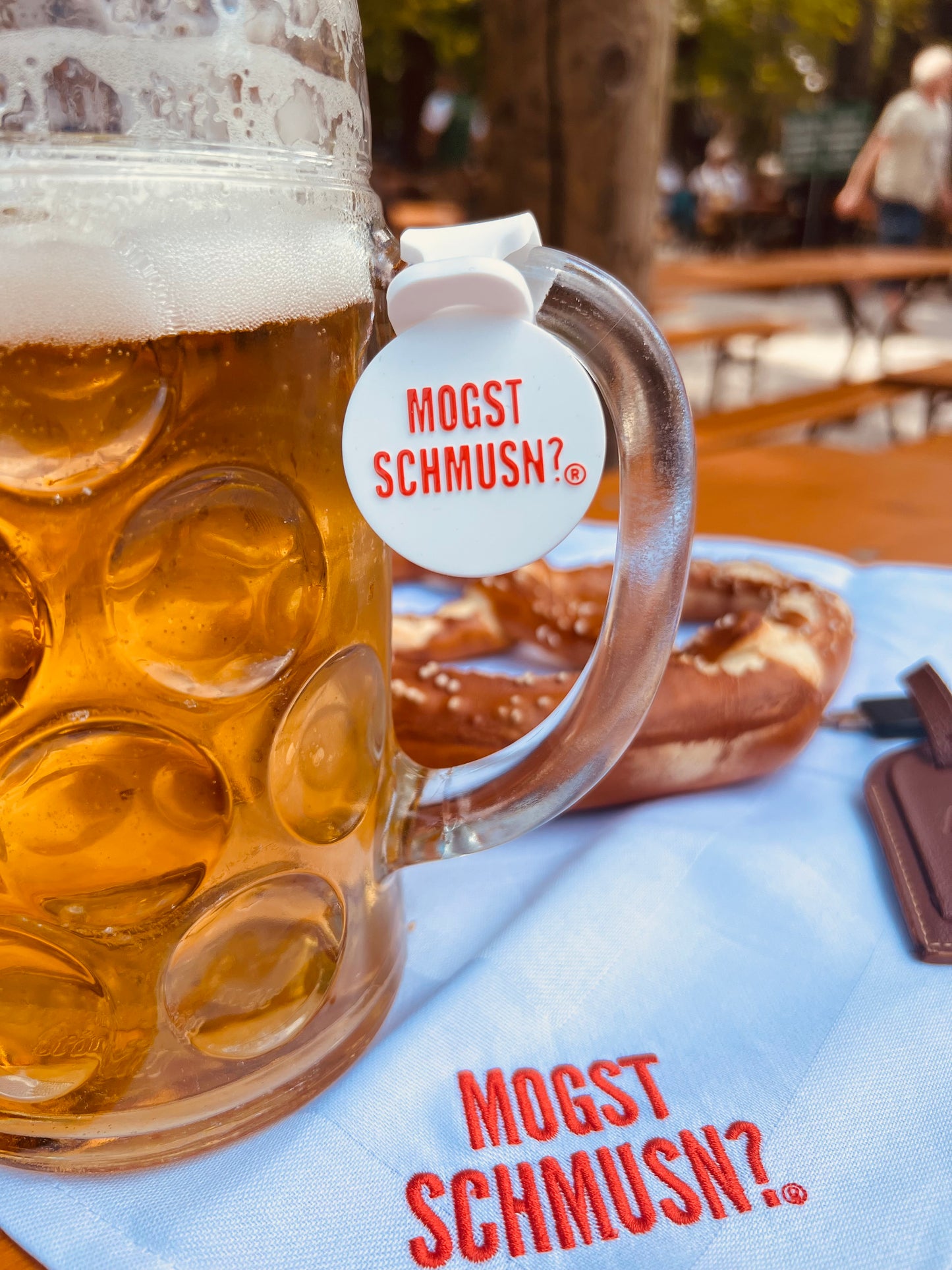 Maßkrug mit goldenem Bier im Biergarten, am Henkel befestigt ein weißes Silikon-Maßkrugbandl mit rotem Aufdruck MOGST SCHMUSN?®. Daneben eine Brezn und ein Tuch mit dem gleichen Schriftzug – stimmungsvolles Wiesn-Accessoire.