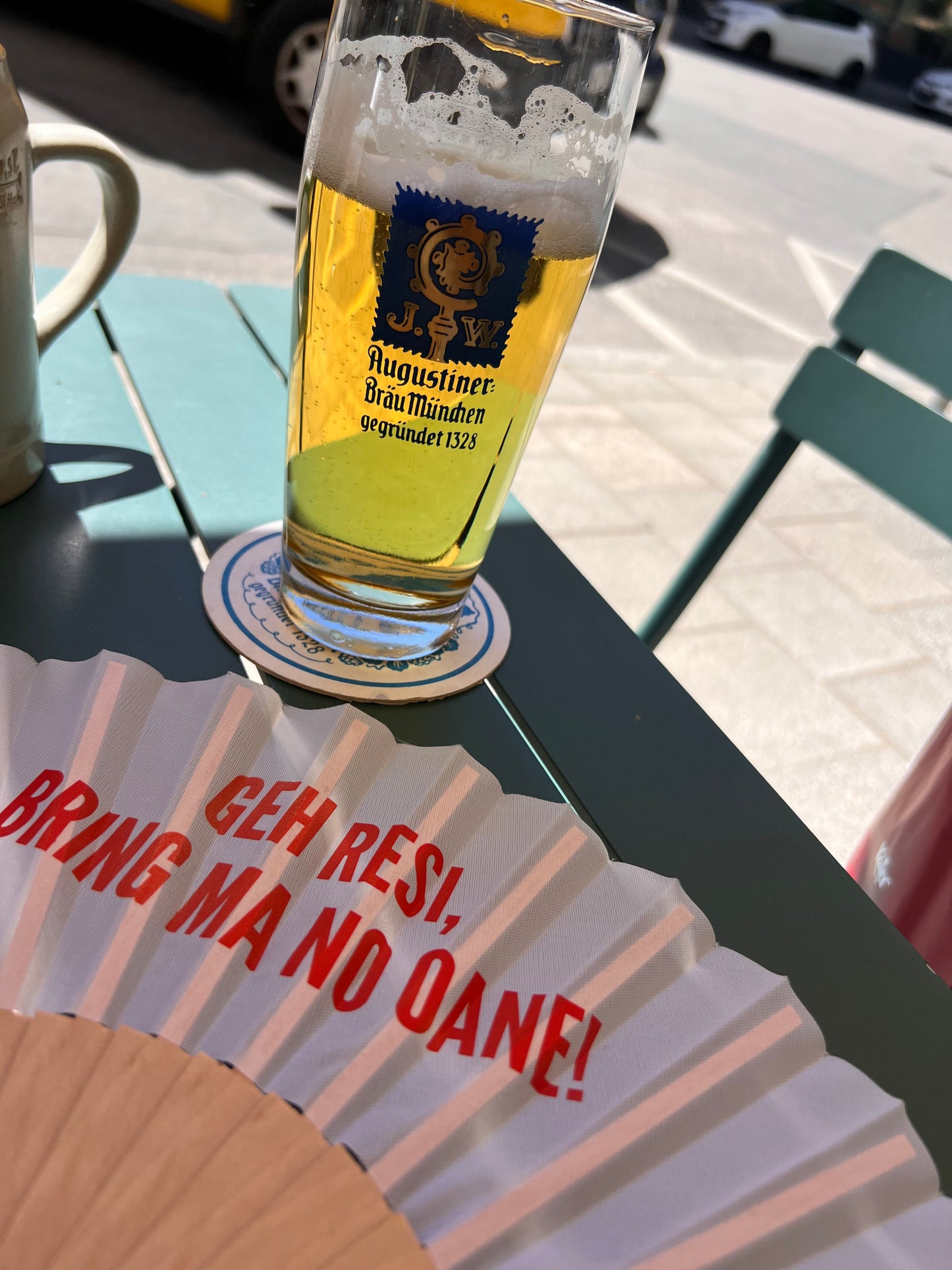 Weißer Fächer mit Aufdruck „Geh Resi bring ma no oane“ neben einem Bierglas im Münchner Biergarten.