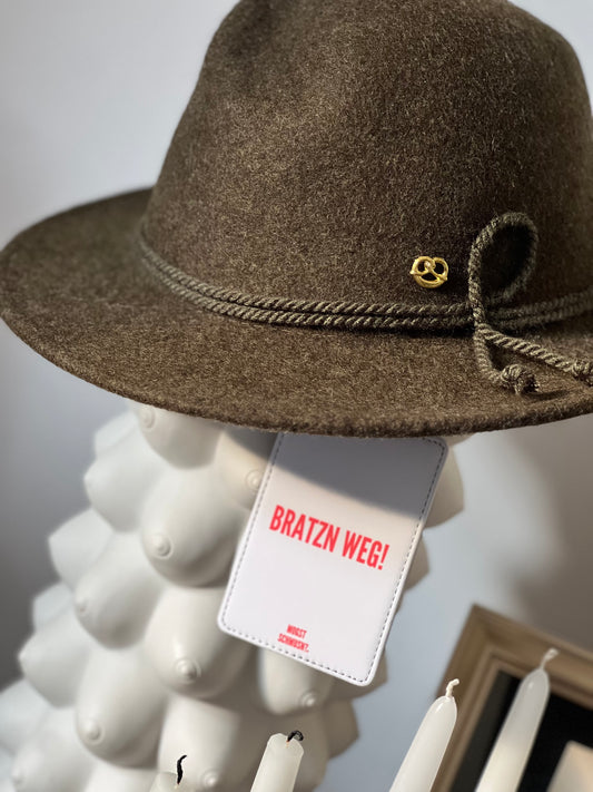 Brauner Trachtenhut mit goldenem Anstecker, daran befestigt ein weißer MOGST SCHMUSN?® Koffer- und Taschenanhänger mit rotem Schriftzug „BRATZN WEG!“ in stilvoller Indoor-Szene.