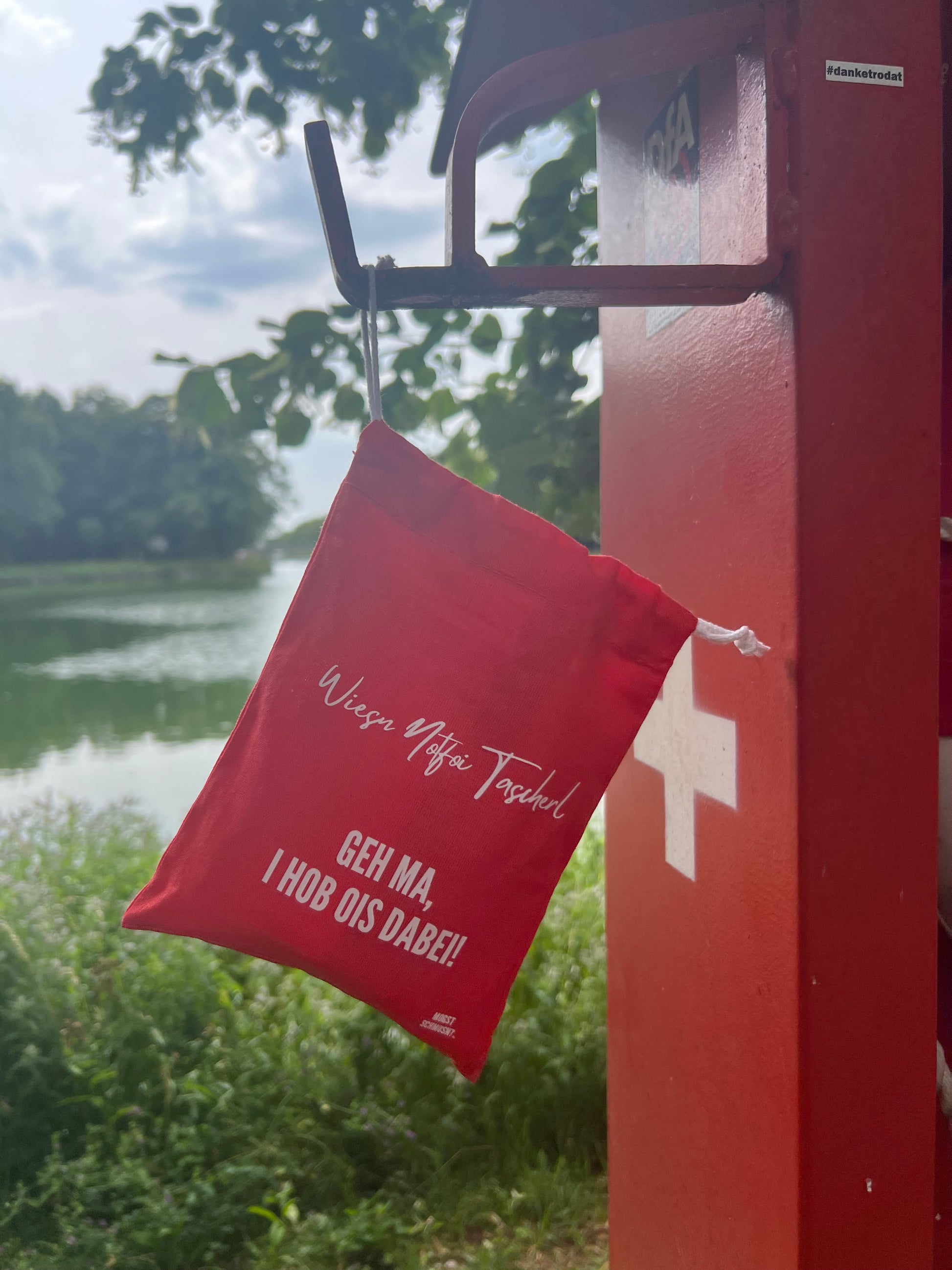 Rotes MOGST SCHMUSN?® Wiesn Notfoi Tascherl mit weißer Aufschrift am Notfallring aufgehängt in Nymphenburg beim Hubertusbrunnen mit Blick auf dem Kanal