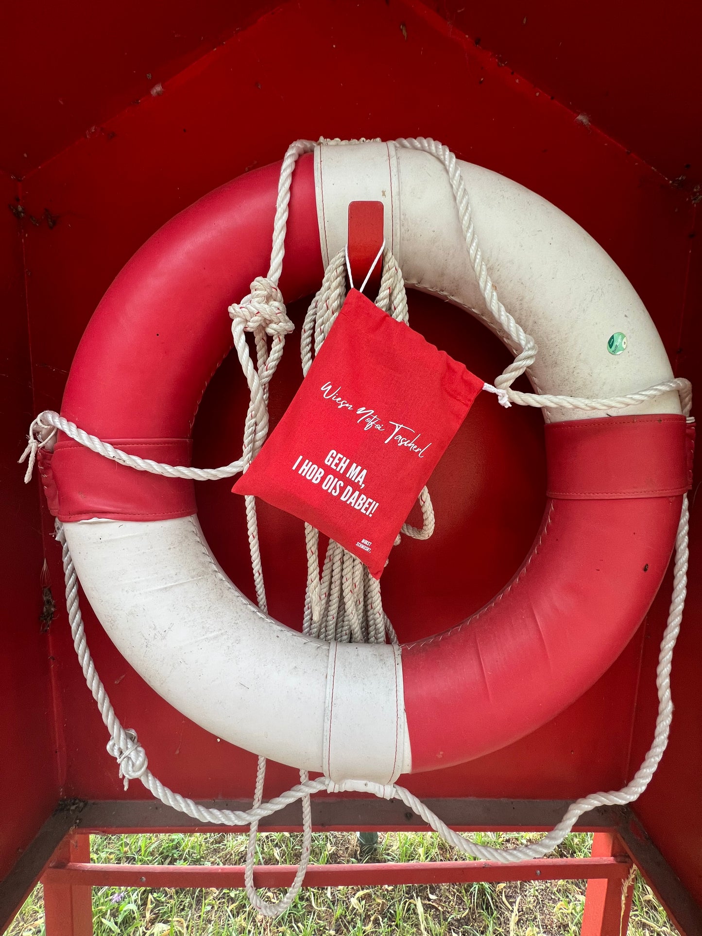 Rotes MOGST SCHMUSN?® Wiesn Notfoi Tascherl mit weißer Aufschrift am Notfallring aufgehängt in Nymphenburg beim Hubertusbrunnen mit Blick auf dem Kanal