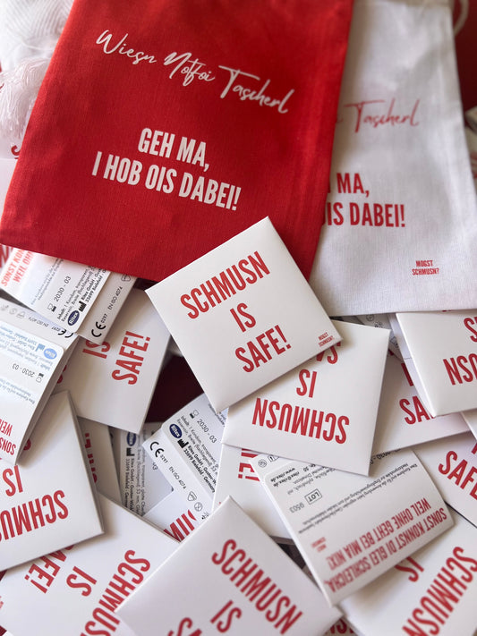 Mehrere weiße Kondomverpackungen mit rotem Aufdruck „SCHMUSN IS SAFE!“ liegen locker verstreut auf einer Oberfläche. Darüber liegen zwei Stofftaschen in Rot und Weiß mit der Aufschrift „Wiesn Notfoi Tascherl – Geh ma, i hob ois dabei!“.