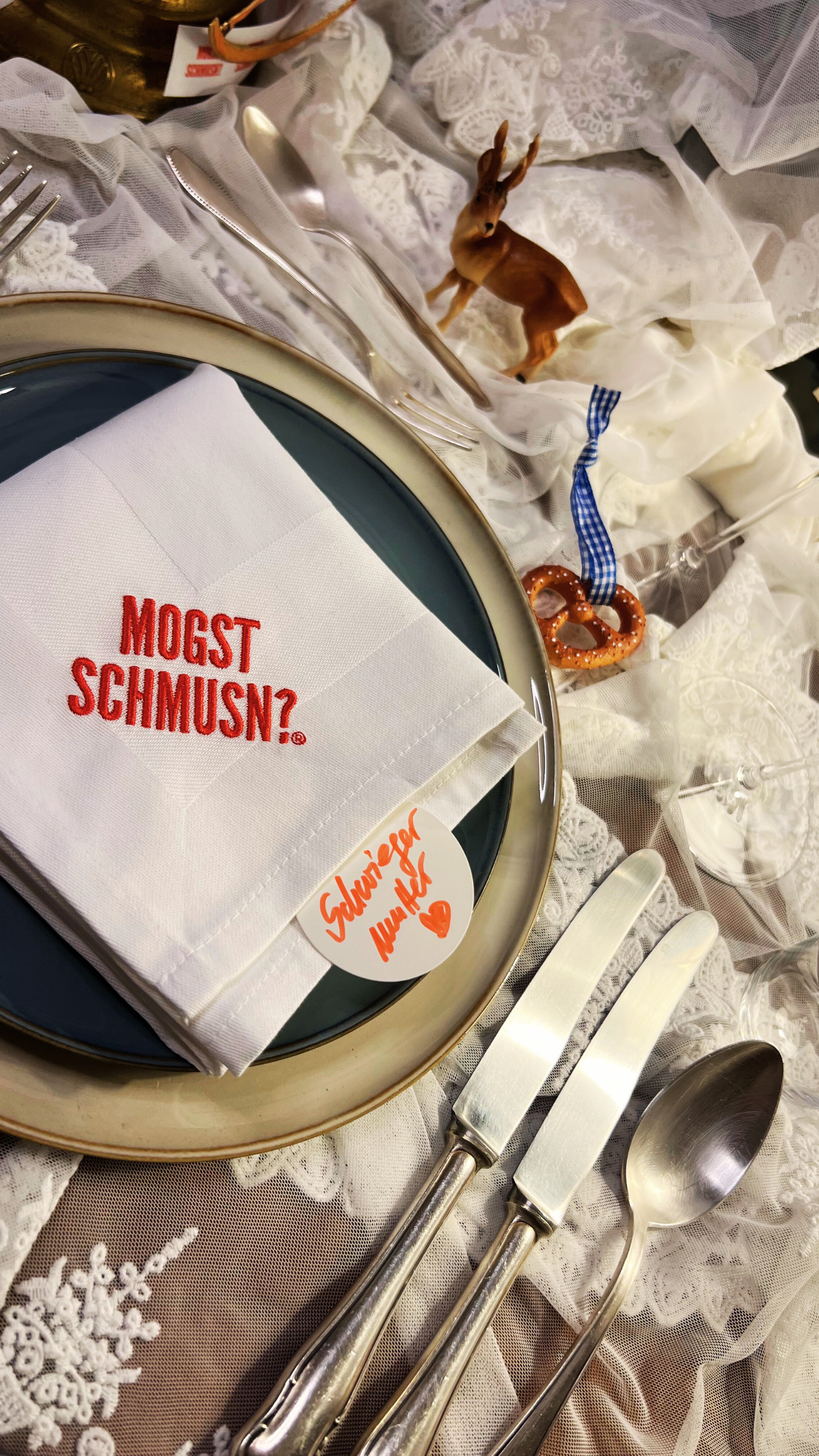 man sieht hier einen festlich gedeckten tisch mit einer organza tischdecke, verschiedene kleine dekoelemente, wie ein elch eine brezn, besteck, zwei teller und eine stoffserviette gefaltet in weiß mit dem Markenlogo MOGST SCHMUSN?® aufgestickt - seitlich ragt ein rundes Namensschild auf dem mir roter Schrift "Schwiegermutter" und ein gemaltes Herz drauf steht.