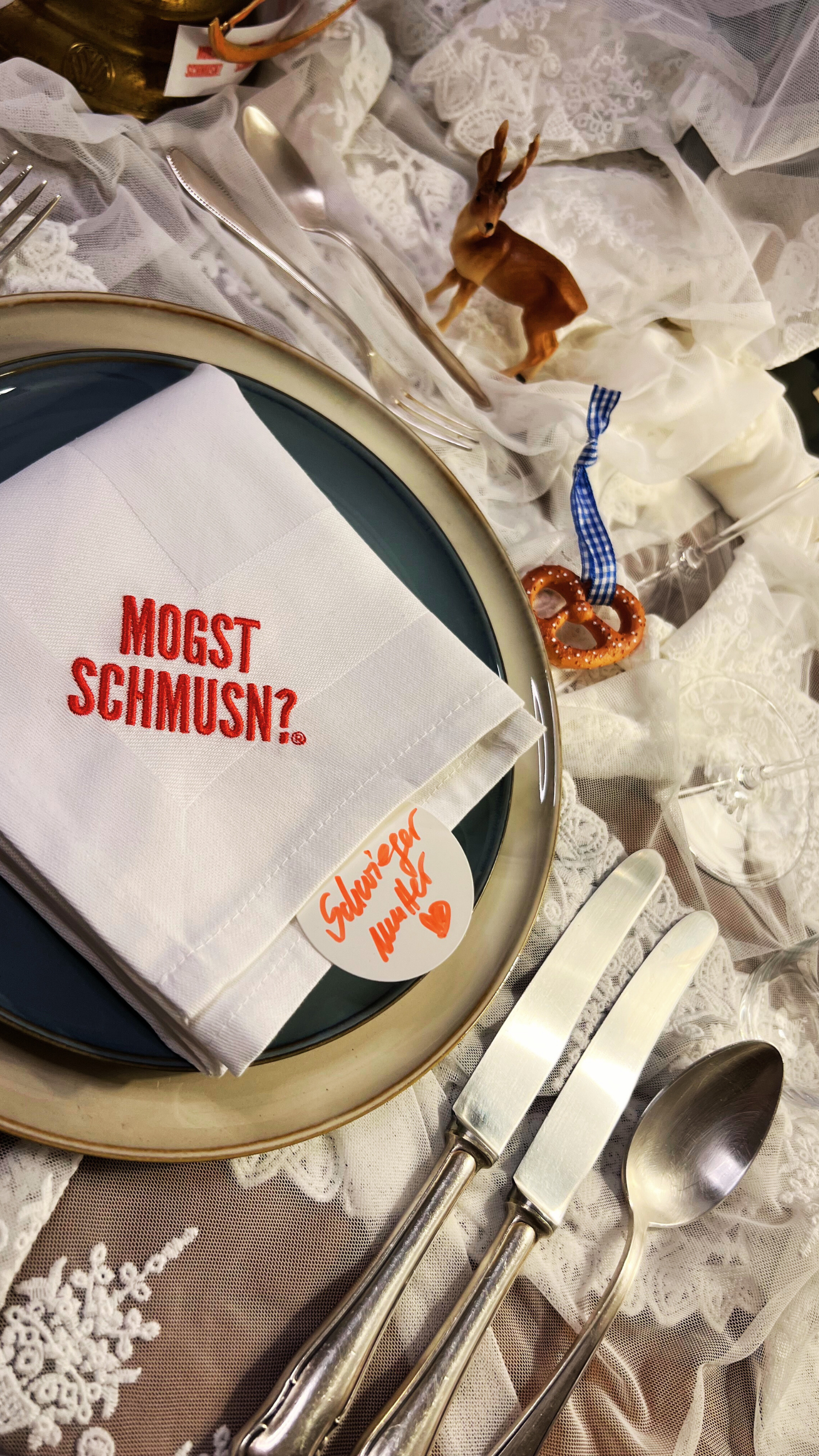 man sieht hier einen festlich gedeckten tisch mit einer organza tischdecke, verschiedene kleine dekoelemente, wie ein elch eine brezn, besteck, zwei teller und eine stoffserviette gefaltet in weiß mit dem Markenlogo MOGST SCHMUSN?® aufgestickt - seitlich ragt ein rundes Namensschild auf dem mir roter Schrift "Schwiegermutter" und ein gemaltes Herz drauf steht.