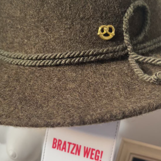 Video vom Brauner Trachtenhut mit goldenem Anstecker, daran befestigt ein weißer MOGST SCHMUSN?® Koffer- und Taschenanhänger mit rotem Schriftzug „BRATZN WEG!“ in stilvoller Indoor-Szene schwenkend zu einem Kerzenständer mit 4 weiße Spitzkerzen mit bayerischer Raute, Stammtisch Wirtshaus Bild im HIntergrund