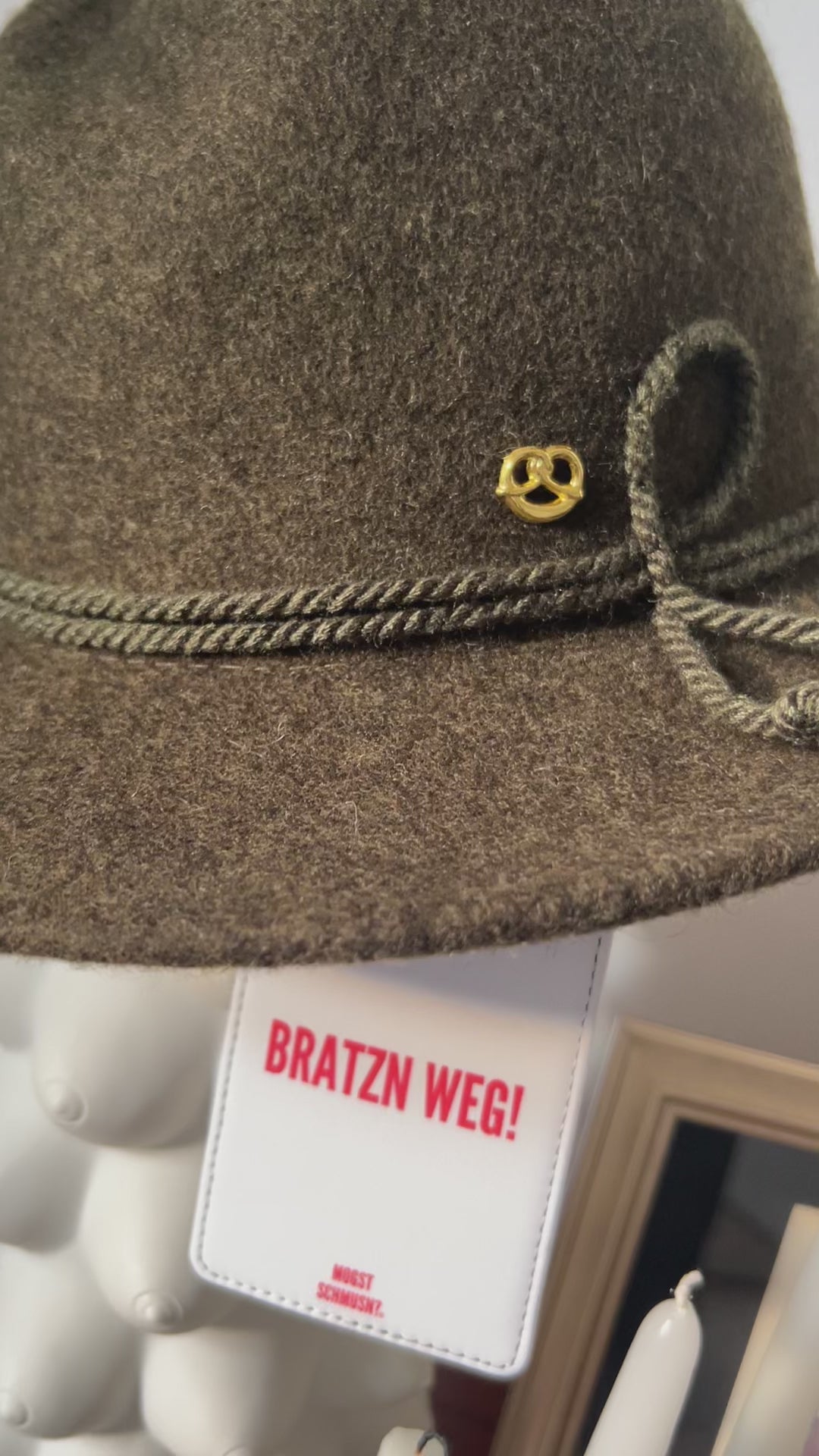 Video vom Brauner Trachtenhut mit goldenem Anstecker, daran befestigt ein weißer MOGST SCHMUSN?® Koffer- und Taschenanhänger mit rotem Schriftzug „BRATZN WEG!“ in stilvoller Indoor-Szene schwenkend zu einem Kerzenständer mit 4 weiße Spitzkerzen mit bayerischer Raute, Stammtisch Wirtshaus Bild im HIntergrund