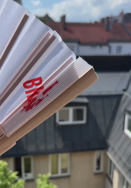 Video mit weißem Fächer mit rotem Aufdruck vor einem geöffneten Fenster im Sonnenlicht.